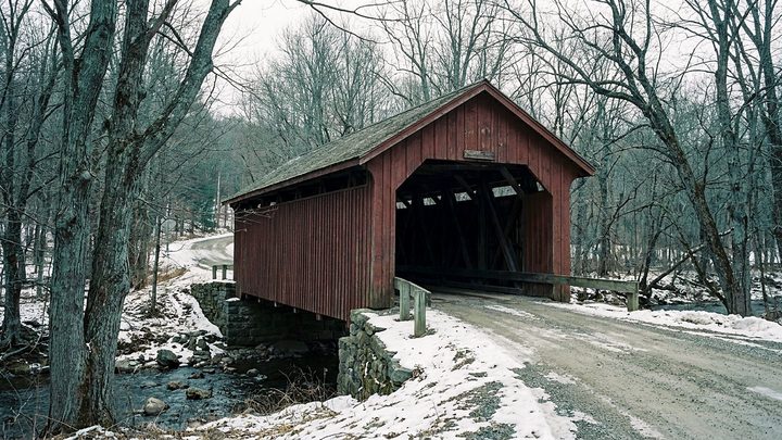 Illustration of a red covered bridge on a snowy back road, its dark opening showing the timber frame above a small stream