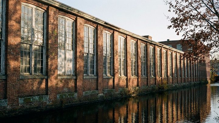 Illustration of a long brick mill wall with tall arched windows above a still canal, lit low by late autumn sun