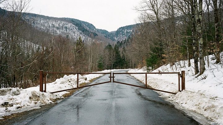 Illustration of a rusted gate closed across a wet mountain road, with snowbanks on both sides and a notch in the ridge beyond
