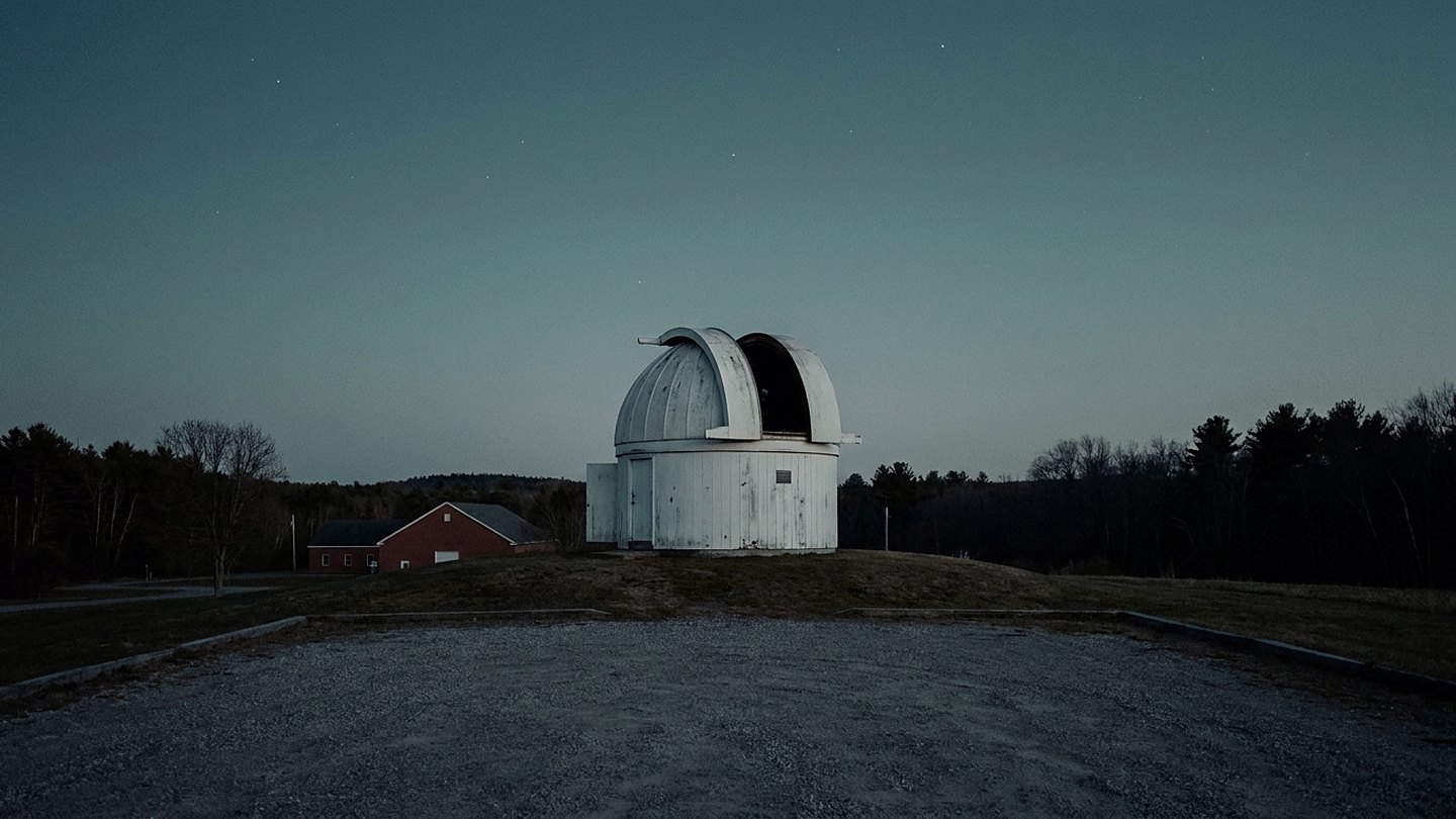 Illustration of a small white observatory dome with its shutter open on a rise, seen from a gravel lot under a deep blue dusk sky